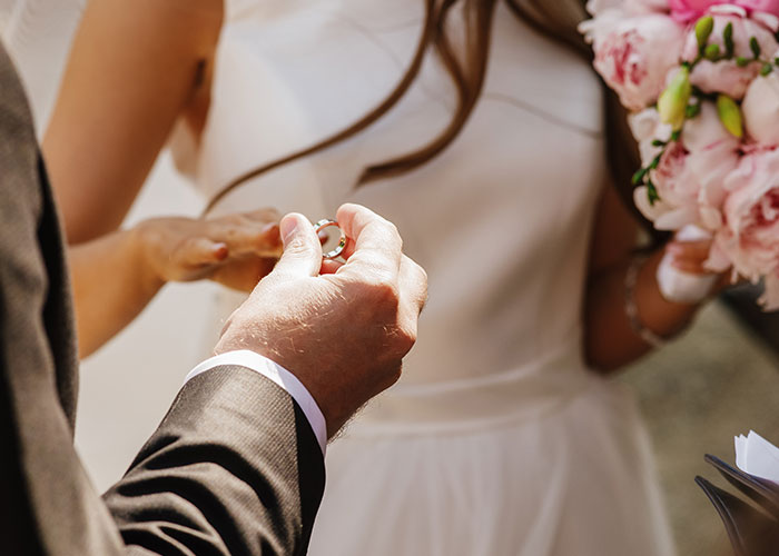 Bride and groom exchanging rings during wedding ceremony, holding bouquet of pink flowers.