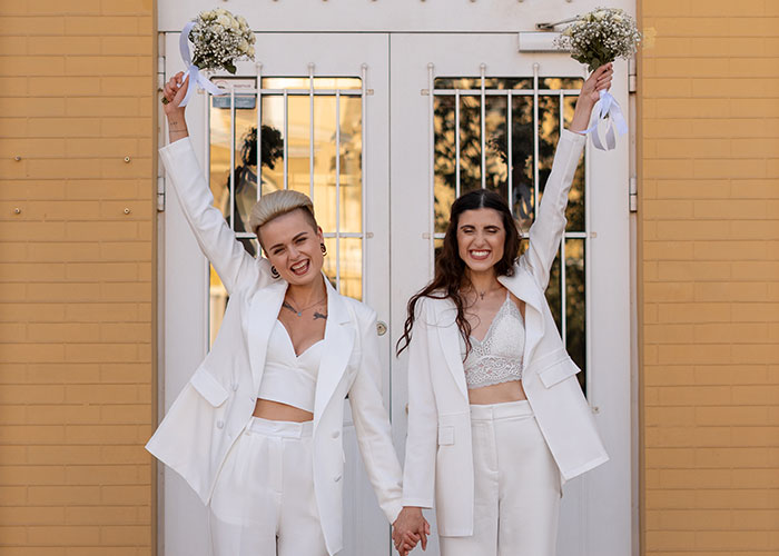 Two women in white suits, holding hands and bouquets, smiling in front of a door, reflecting partner views.