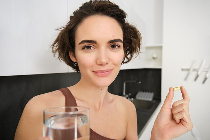 Woman demonstrating a crazy body life hack by holding a water glass and a capsule in a modern kitchen.