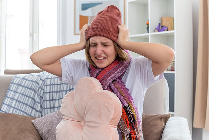 Person with a headache holding their head, using a warm hat and scarf indoors as a crazy body life hack.