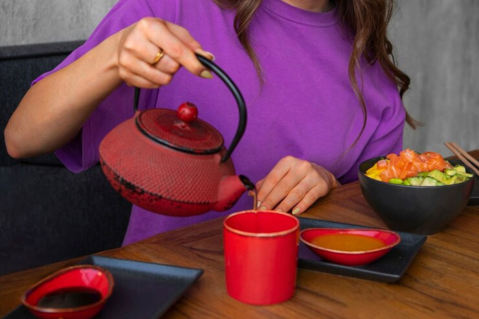 Woman pouring tea from a red teapot into a matching cup, illustrating crazy body life hacks over a meal.