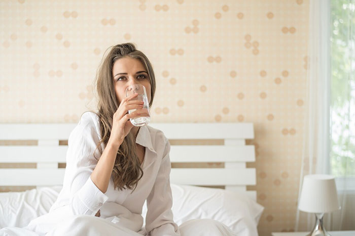 Woman in a white shirt drinking water in a bedroom, illustrating a life hack.