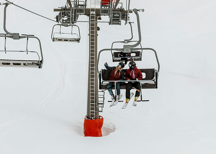 Ski lift with two people seated, wearing ski gear, set against a snowy landscape, capturing a candid winter adventure moment.