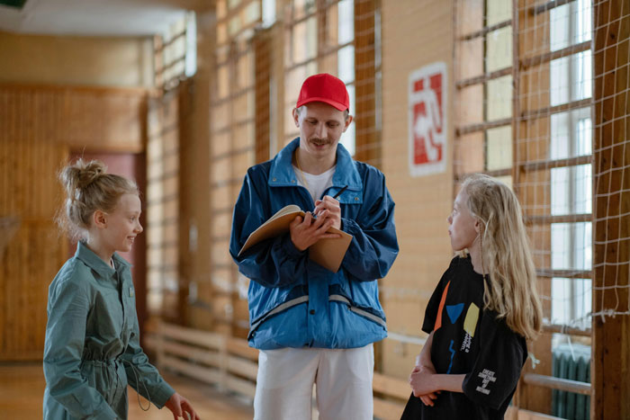 Man with a red cap and clipboard coaching two young girls in a gym.