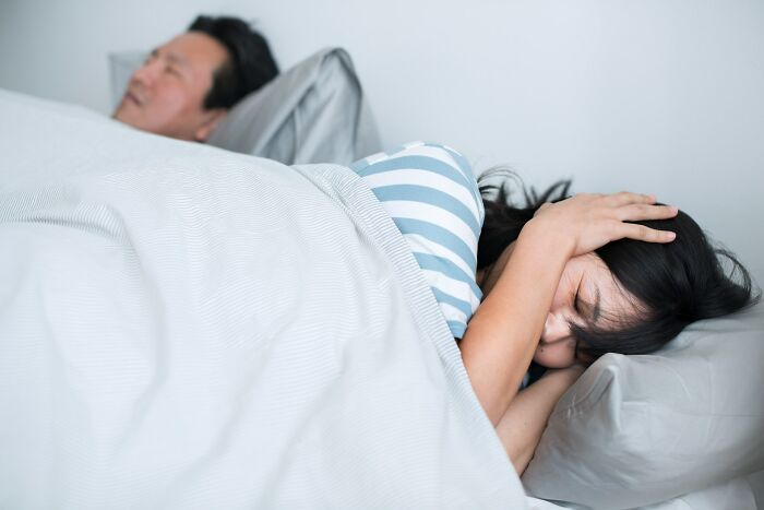 Woman covering ears in bed, man sleeping beside her; focuses on health advice about the benefits of exercise.