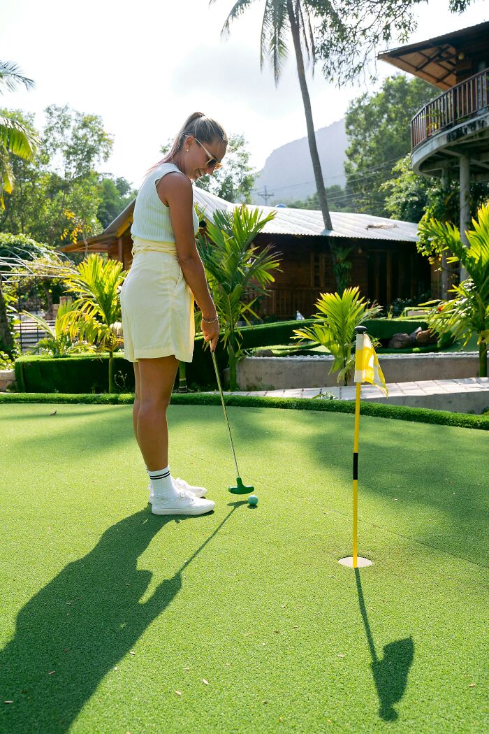 A woman playing mini-golf on a sunny day with palm trees and a wooden cabin in the background.