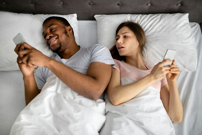 Couple lying in bed, both using phones, conveying secrets in relationships.