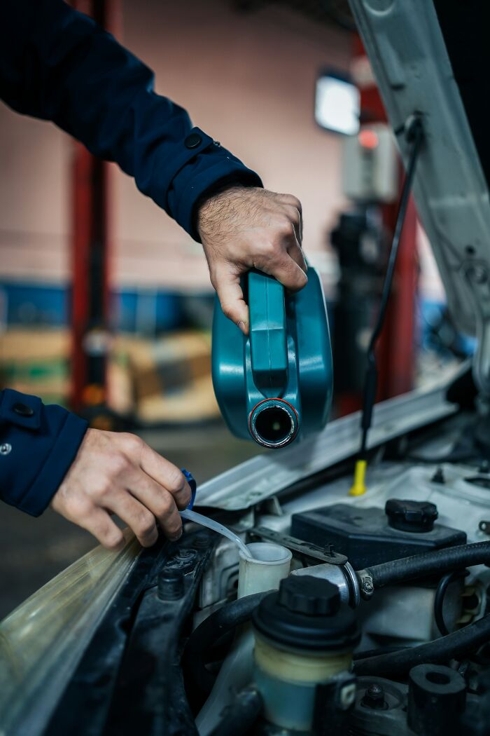 Person pouring oil into a car engine, wearing a blue jacket, in a garage setting, connected to secrets from loved ones.