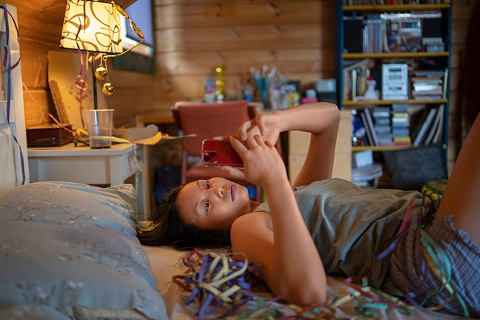 Teen lying on bed using phone, surrounded by clutter in a cozy room environment, illustrating struggles with control.