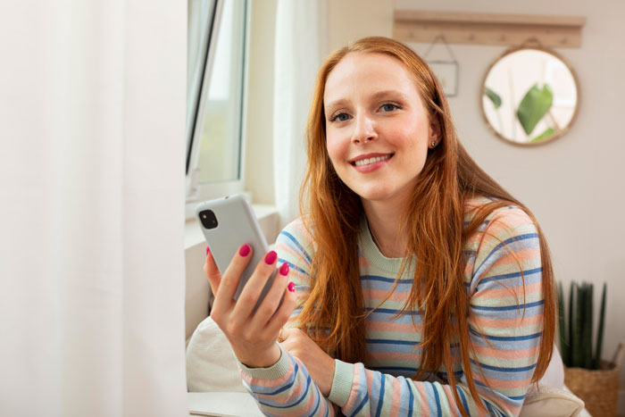 Young woman in a striped sweater smiling, holding a phone, symbolizing a daughter using a mom tracker app.