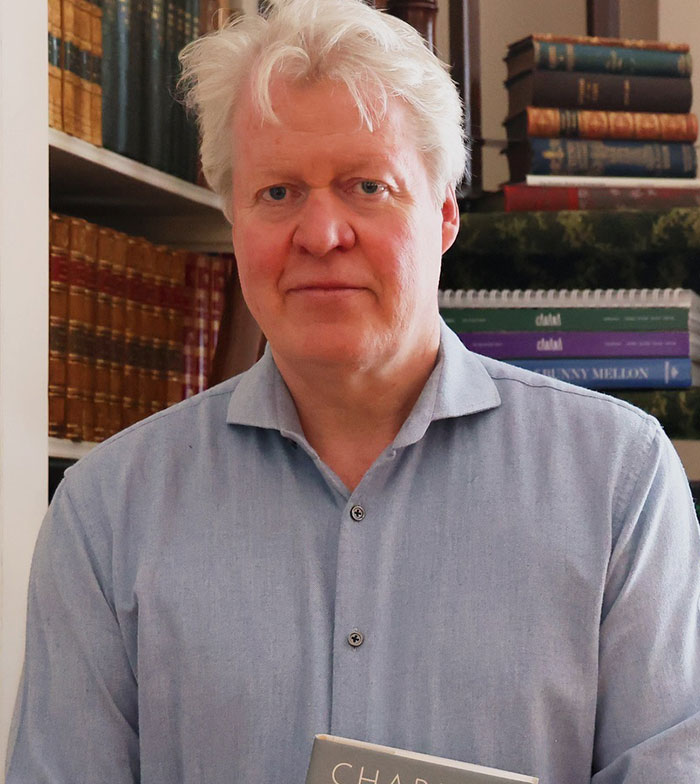 An individual stands in a library, holding a book. Shelves filled with stacked books are visible in the background.