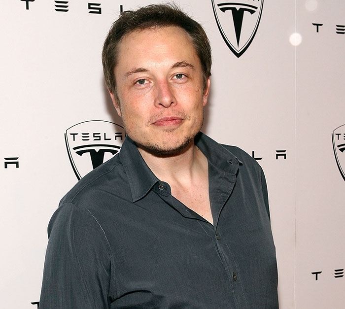 Younger Elon Musk in front of Tesla logo in a dark shirt