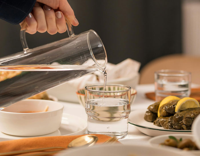 Traveler pouring water into a glass at a dining table, featuring stuffed grape leaves with lemon slices.