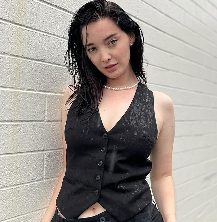 Non-binary person wearing a wet black vest, posing against a white brick wall.