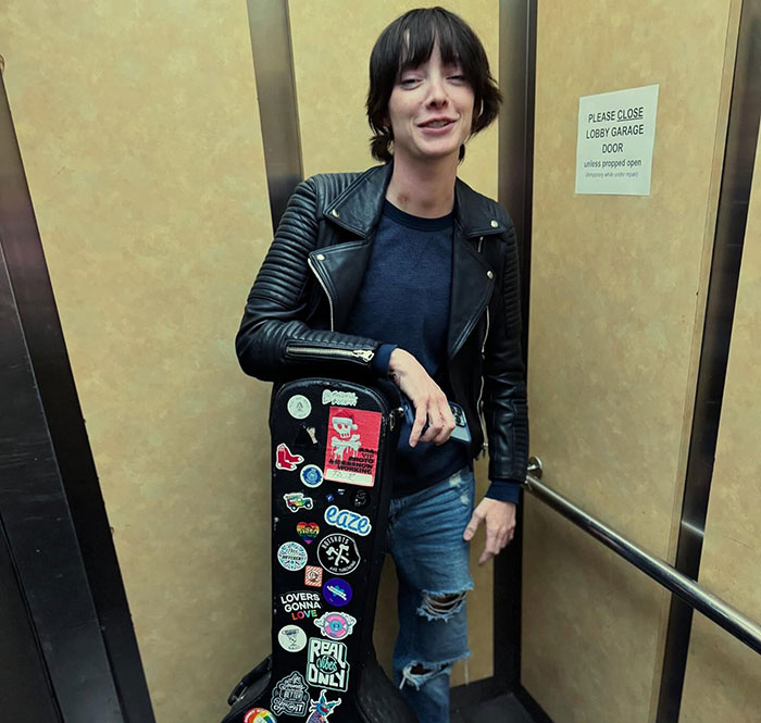 A person in an elevator wearing a leather jacket, holding a sticker-covered guitar case; related to "Oppenheimer" star.