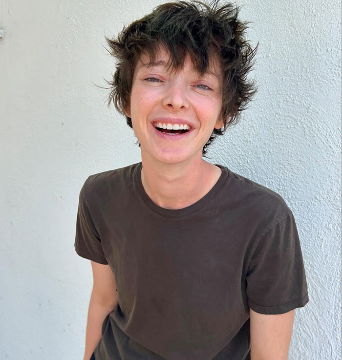 A smiling person in a casual dark t-shirt against a white wall, associated with 'Oppenheimer'.