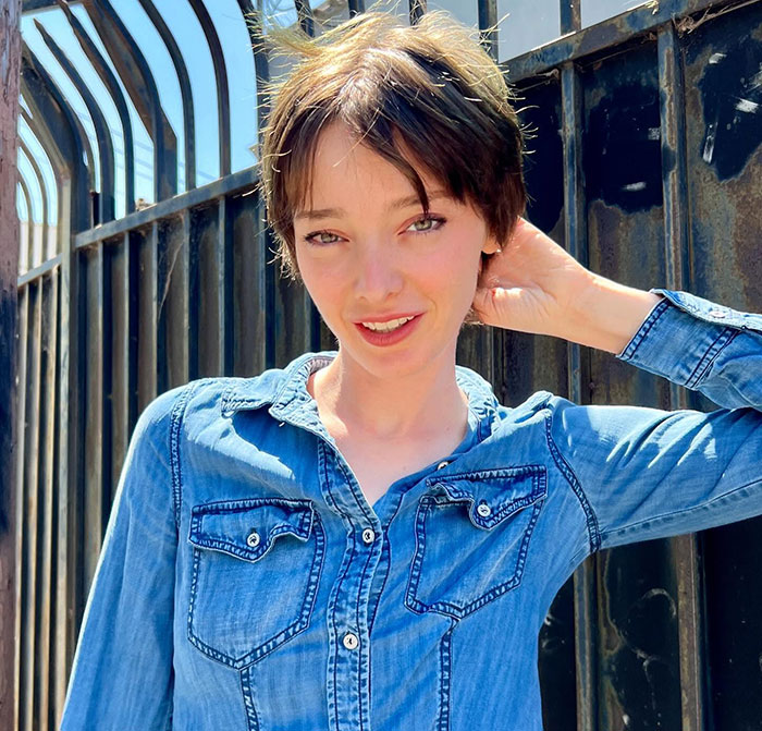 A person outdoors in denim, smiling in front of a metal fence, trans masculine non-binary theme prominent.