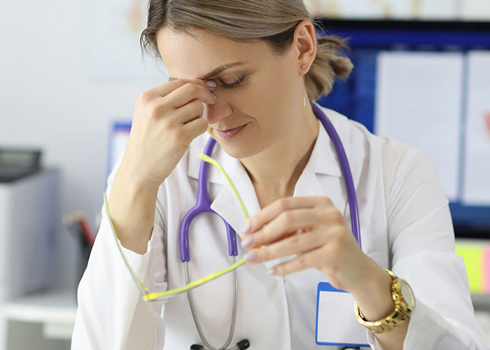 Doctor looking stressed over employer demands on overpayment, holding glasses at a desk.