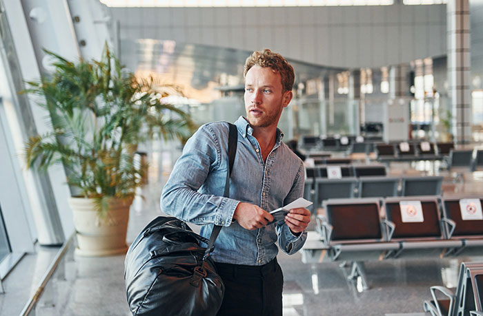 A man in a denim shirt at airport holding a ticket near empty seating area.