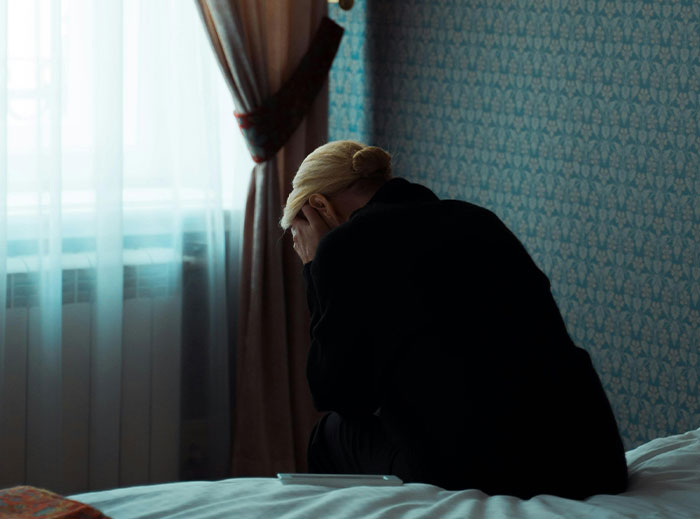 A distressed person sits on a bed, hands covering face, in a dimly lit room next to a window.