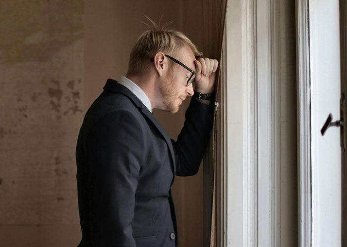 Man in a suit and glasses looking stressed by a window, illustrating CEO management challenges.