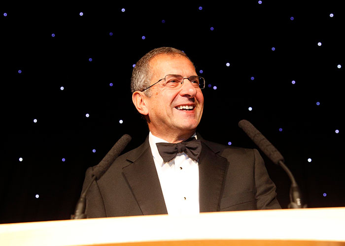 A smiling man in a tuxedo stands at a podium under a starry backdrop, representing big CEO management challenges.