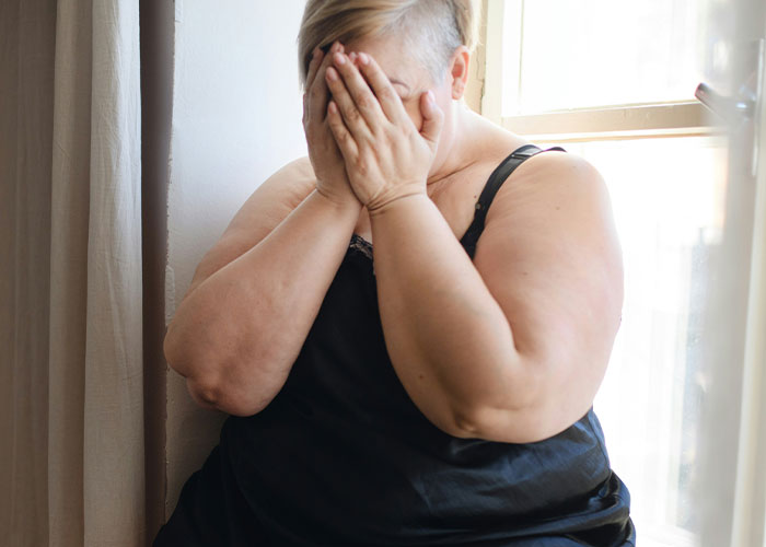 Person in black camisole sitting by a window, covering face, expressing distress about fat-shaming.