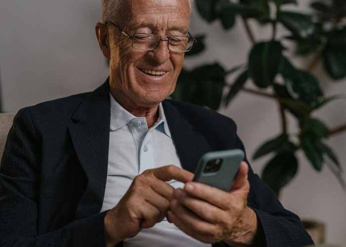 Elderly man smiling at smartphone, wearing a dark blazer and glasses, with plants in the background.