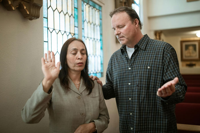 A woman and man in thoughtful prayer, holding hands, in a church setting with stained glass windows.