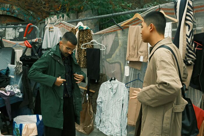 Two men at an outdoor market, one trying on a green coat, while surrounded by various clothing items, silently judged by style.