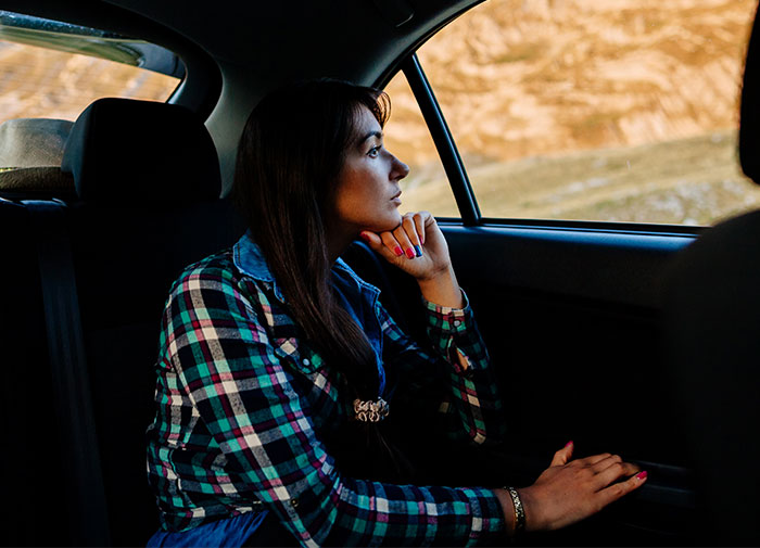 Woman in a checkered shirt sitting thoughtfully in a car, looking out the window, relating to a fiance demanding to sell the car.