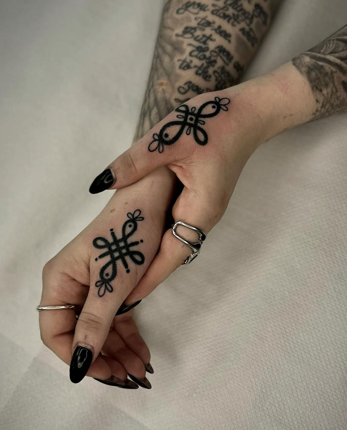 Unique black ink finger tattoo designs on hands with rings and dark nails.