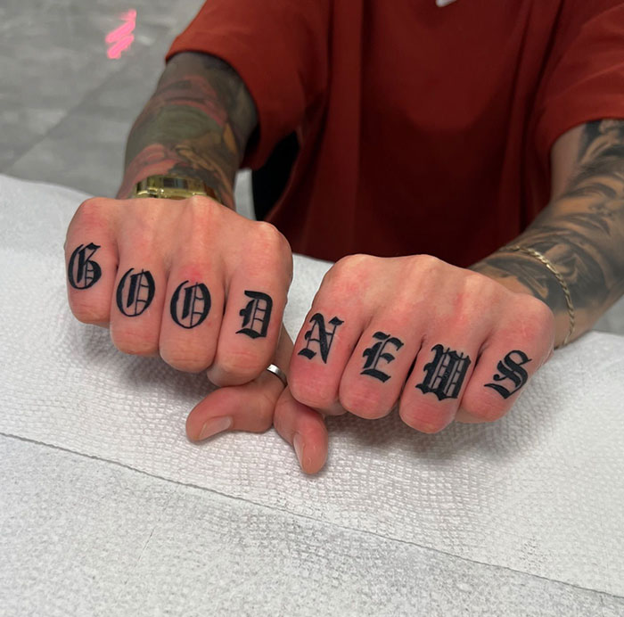 Tattooed knuckles displaying "GOOD NEWS" in bold font, showcasing finger tattoos.