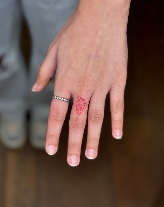 Hand with finger tattoos featuring a small red anatomical heart design, worn with a silver ring.