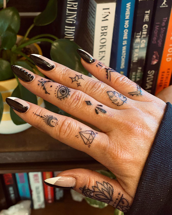 Hand with intricate finger tattoos and black nails against a background of books.