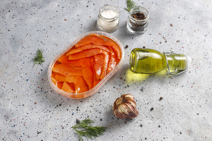 Smoked salmon in a plastic container with garlic, oil, and spices on a speckled surface.