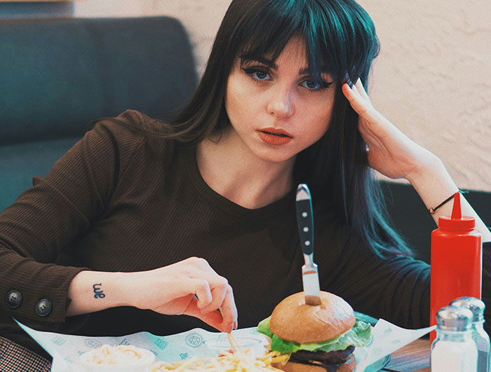 Woman sitting at a table with a hamburger and fries, looking frustrated, highlighting friend always late issue.