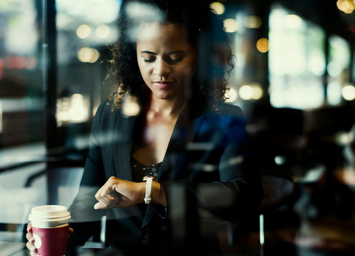 Woman checking watch, holding coffee cup, seemingly frustrated by friend always being late.