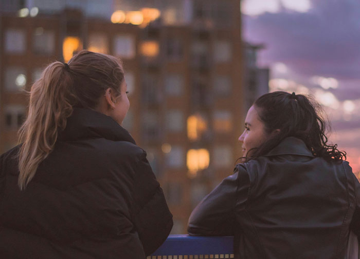 Two friends in jackets chatting on a balcony at sunset with urban buildings in the background.