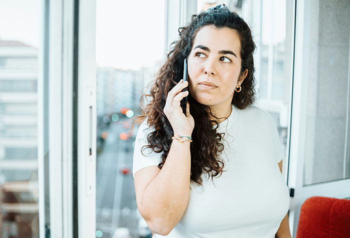 Woman on phone, thoughtful expression, by a window, discussing a friend's delayed repayment.