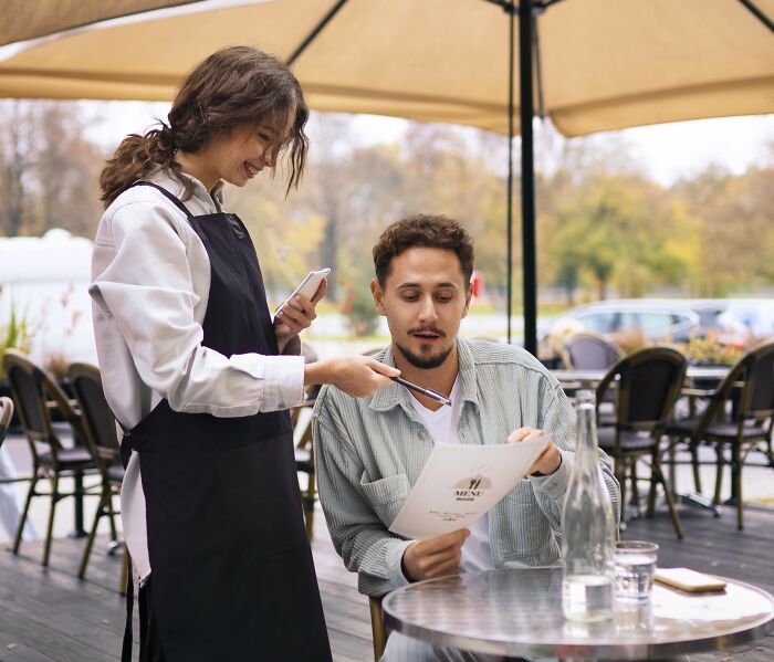 Traveler looks confused with menu while ordering from smiling waitress at outdoor cafe.