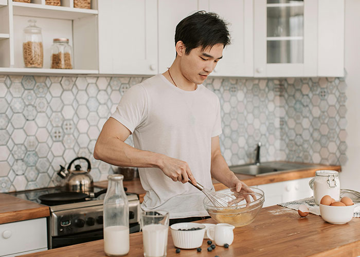 Man in a kitchen, using frugal hacks to whisk eggs in a bowl, with milk and blueberries on the counter.