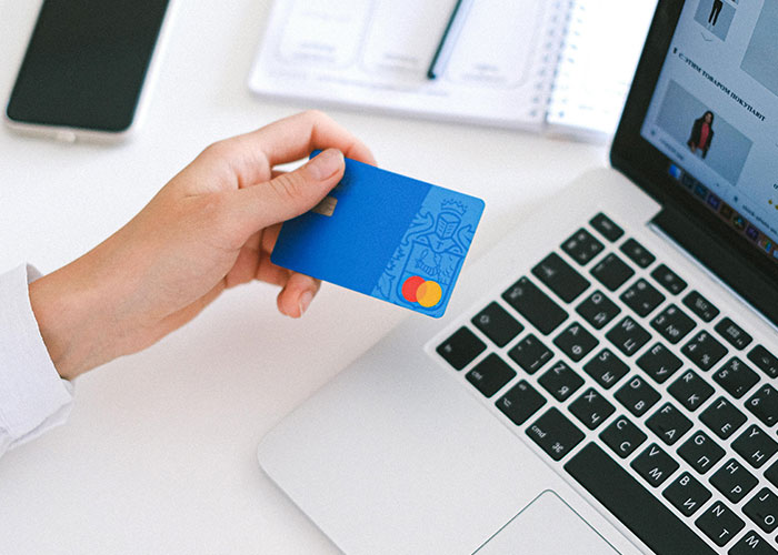 Person holding a blue credit card near a laptop, illustrating frugal hacks for saving money.