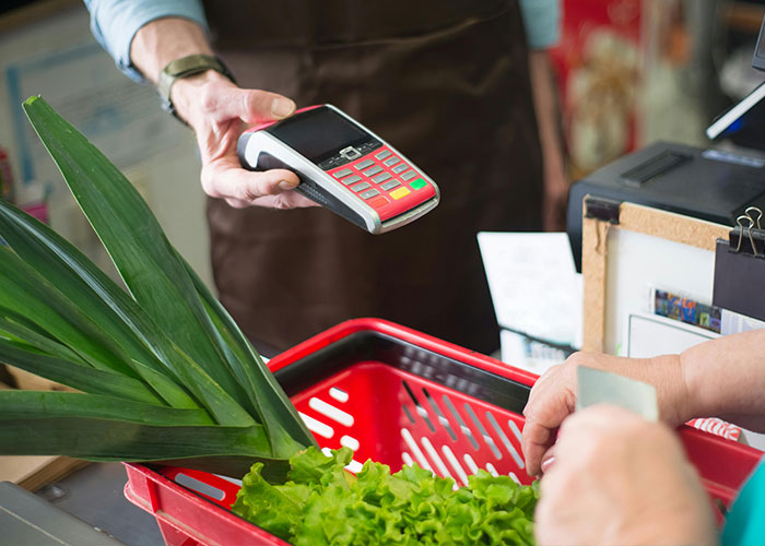 A person using a card reader for payment over a basket of fresh produce, demonstrating frugal hacks in a grocery store.