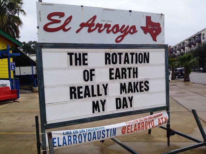 El Arroyo sign with humorous message about Earth's rotation in a street setting.