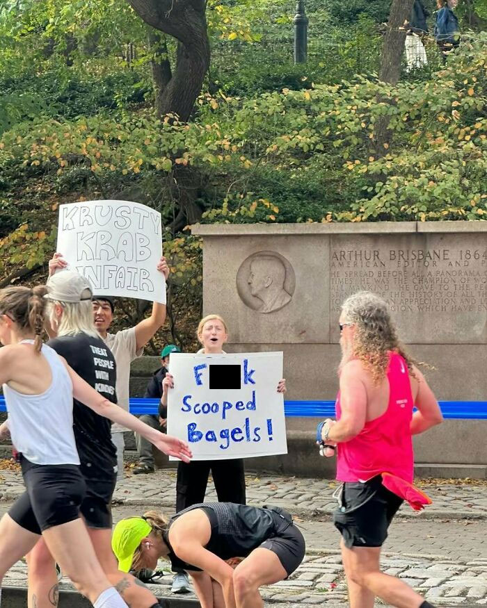 People in a New York park holding humorous protest signs, including one about scooped bagels.