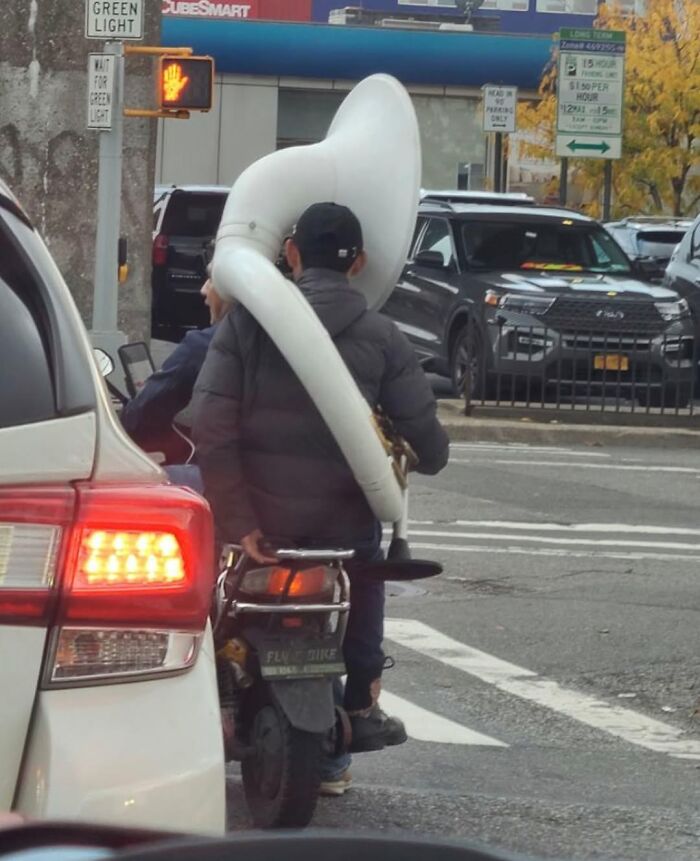 Man on scooter in New York carrying a large tuba, showcasing humorous street scene.