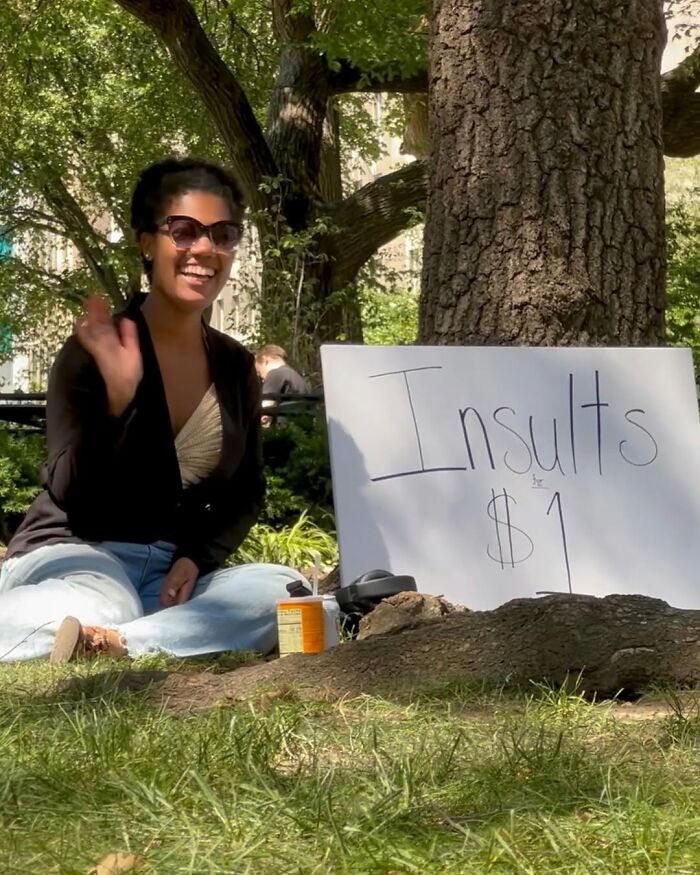 Person sitting in a park in New York, smiling beside a sign offering insults for $1.