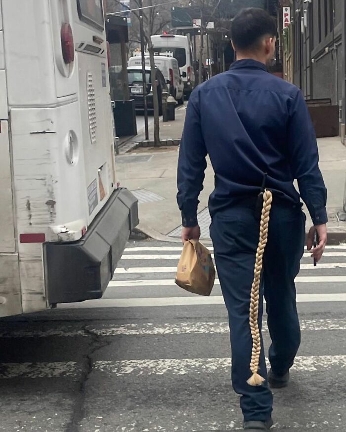 Man in blue uniform with a long braided ponytail attachment walking in New York street.