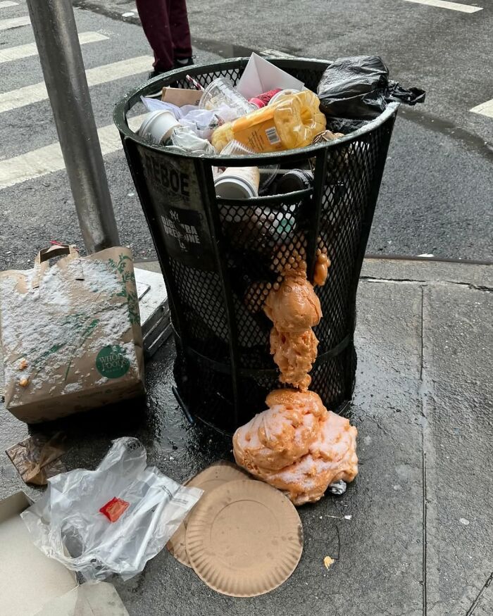 Overflowing trash can on a New York street corner, with food waste spilling onto the sidewalk.